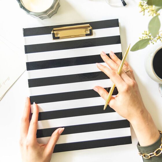 hands holding black and white striped clipfolio, gold clip and pen, coffee, candle, dried leaves