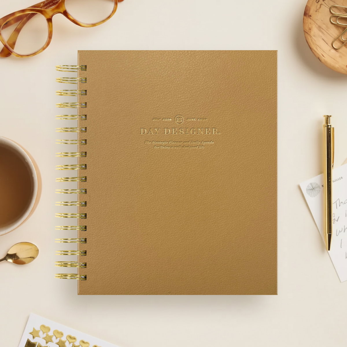 caramel colored day designer planner, gold binding and pen, coffee, glasses, dish with paperclips, gold stickers, beige background
