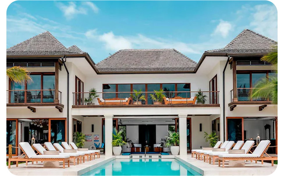 Two‑storey home with a pool, patio seating, umbrellas, and balconies under a bright sky.