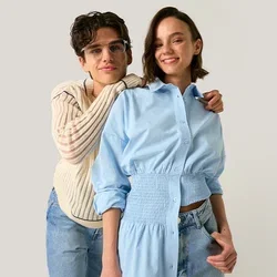 Deux jeunes femmes se tiennent proches, l'une porte des lunettes et un pull crème et embrasse une autre vêtue d'une robe chemise bleue, toutes deux souriantes sur un fond uni.