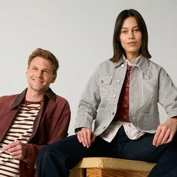 Homme appuyé sur une caisse en bois et femme assise dessus, tous deux portant des vestes décontractées et des pantalons foncés, sur un fond clair uni.