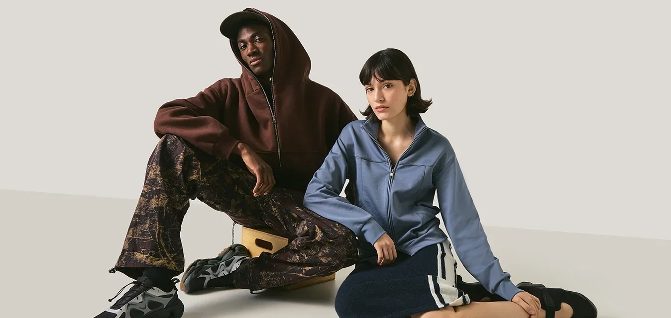 Deux personnes assises sur un sol simple devant un fond neutre, l'une portant un sweat à capuche et un pantalon à motifs, l'autre une veste bleue et une jupe.