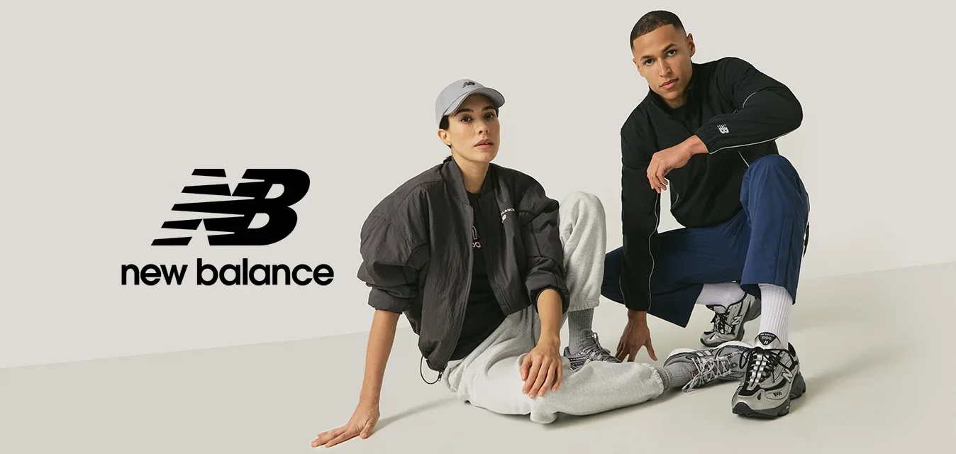 Une femme en casquette grise, veste noire et pantalon de jogging gris est assise à côté d'un homme en veste noire et pantalon bleu, tous deux portant des baskets grises sur un fond uni.