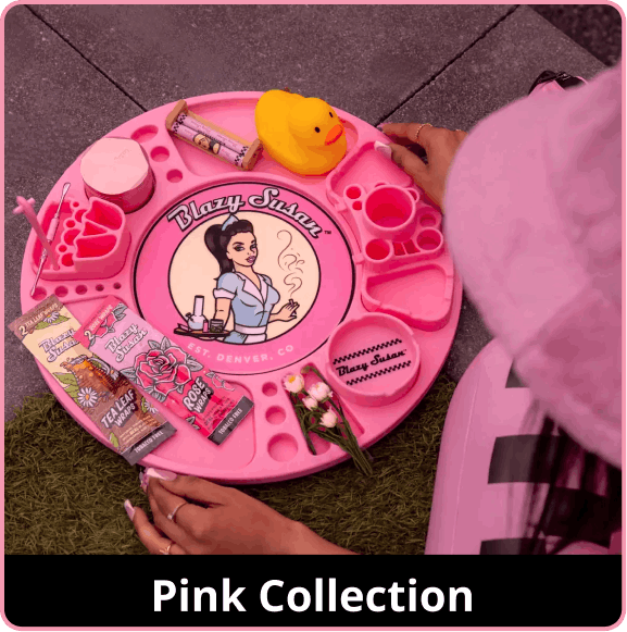 An overhead shot of a large, bright pink circular Blazy Susan spinning rolling tray sitting on a patch of grass next to a stone walkway. The tray is organized with a pink herb grinder, a yellow rubber duck, pink 'Rose Wraps', 'Tea Leaf Wraps', and small flowers. A person wearing a pink furry hat and pink clothing is partially visible on the right. A black banner at the bottom reads 'Pink Collection'.