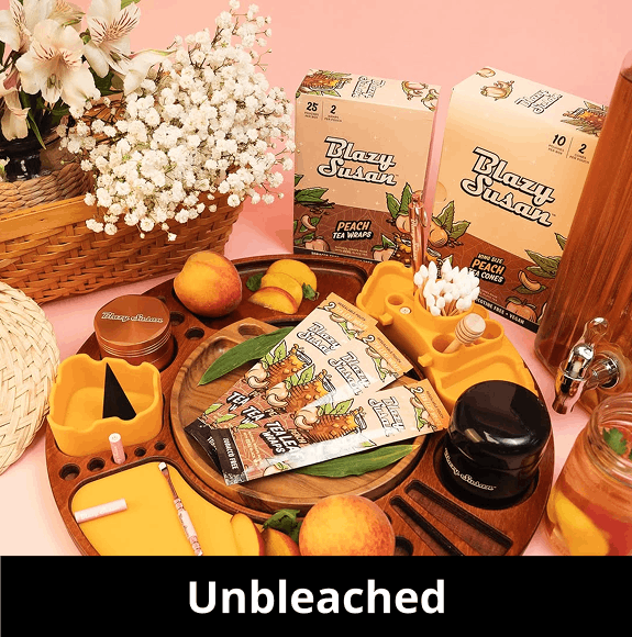A warm, peach-toned flat lay featuring Blazy Susan 'Unbleached' products. In the center is a dark wood circular rolling tray holding several 'Peach Tea Leaf Wraps'. Surrounding the tray are a yellow silicone organizer filled with cotton swabs and a honey dipper, fresh peaches, sliced peaches, a wooden basket of white baby's breath flowers, and boxes of Peach Tea wraps and cones. A black banner at the bottom reads 'Unbleached'.