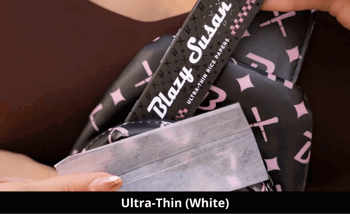 Close-up shot of a person's hands holding an open pack of Blazy Susan 'Ultra-Thin Rice Papers' against a black patterned background. A single, translucent white rolling paper is being held up to demonstrate its thinness. The black packaging features pink cross and diamond patterns with white branding. A black banner at the bottom contains the text 'Ultra-Thin (White)