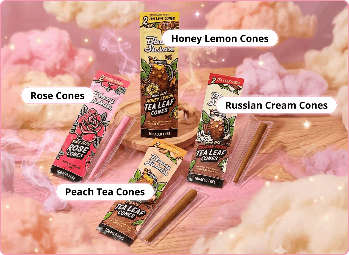 A lifestyle product shot of Blazy Susan's flavored cone collection arranged on a wooden surface surrounded by soft pink and cream cotton clouds. Four distinct packages are labeled with text callouts: 'Honey Lemon Cones' (yellow packaging with a tea pitcher illustration), 'Russian Cream Cones' (green packaging with a creamy drink illustration), 'Rose Cones' (pink packaging with a large red rose), and 'Peach Tea Cones' (tan packaging with a peach and tea illustration). Examples of the pre-rolled cones are shown in clear protective holders next to their respective packs.