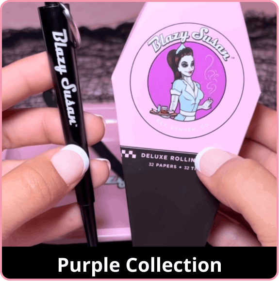 Close-up of hands holding Blazy Susan purple collection items. On the left, a hand holds a black poker tool with white branding. On the right, a hand holds a coffin-shaped 'Deluxe Rolling' pack featuring a purple pin-up girl graphic and a purple circular background. The background is a soft pink. A black banner at the bottom reads 'Purple Collection'.