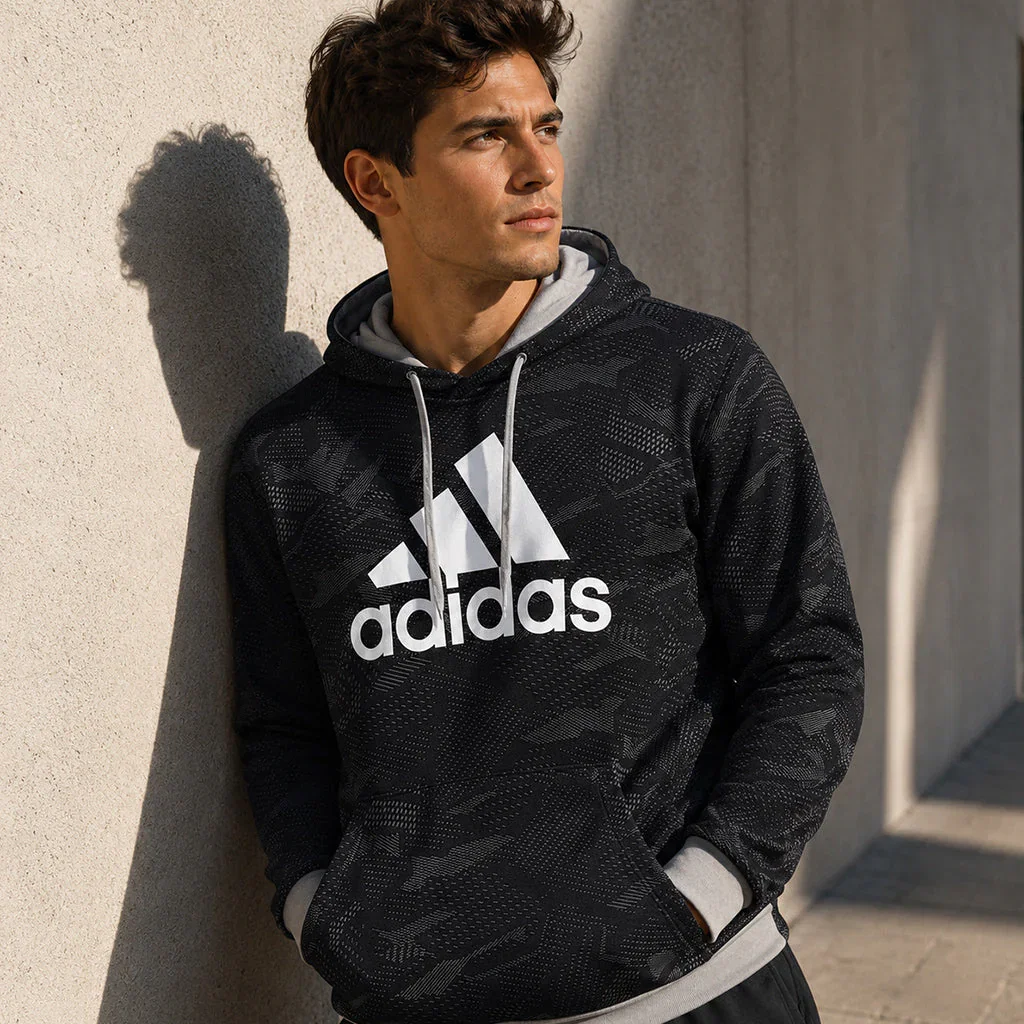 Man wearing black adidas hoodie with white logo and drawstrings, standing against neutral wall in natural sunlight.