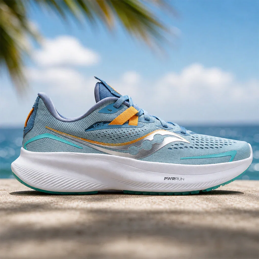 Saucony running shoe in light blue with orange and teal accents displayed on a sandy beach with ocean background.