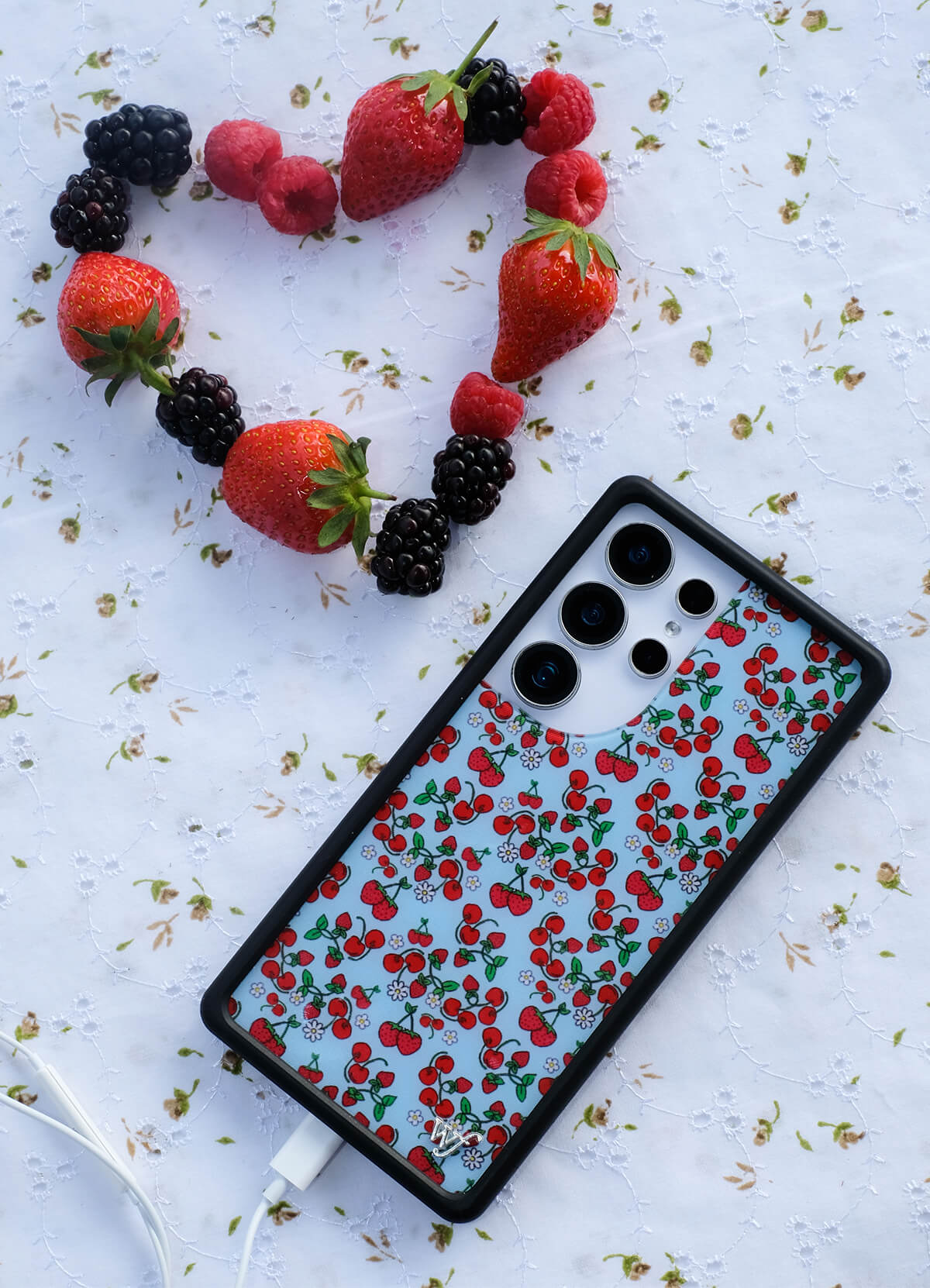 Samsung phone case with a blue background covered in illustrated cherries, strawberries, and daisies, laid on a floral fabric next to a heart shape made from real strawberries, raspberries, and blackberries