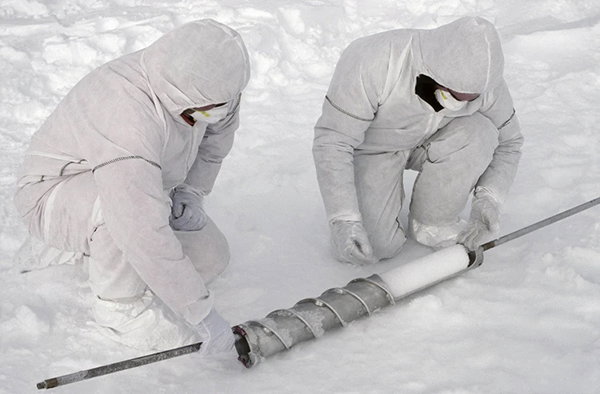 Two researchers wearing white protective coveralls and facemasks kneel in the snow to extract an ice core from a drill bit.