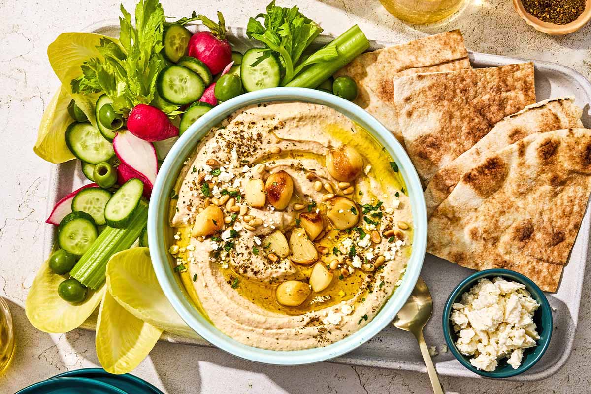 A bowl of roasted garlic hummus on a platter with vegetables, pita wedges and feta cheese.