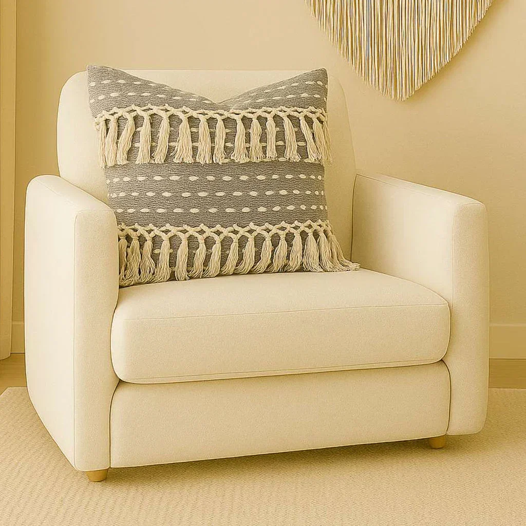 Cream upholstered armchair with tassel-trimmed woven pillow in neutral tones, Reliance Textiles collection.