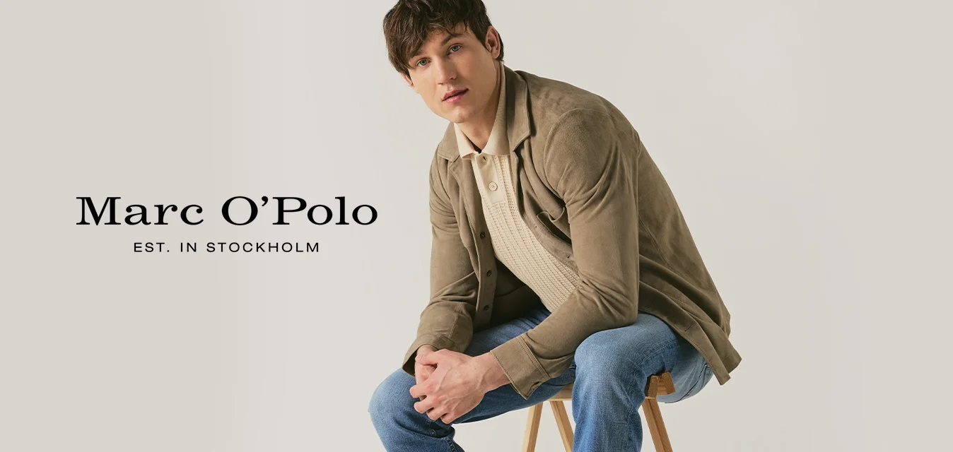 Joven con chaqueta marrón claro, camiseta de punto beige, vaqueros azules y zapatillas blancas sentado en un taburete de madera frente a un fondo neutro.