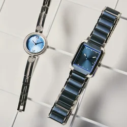Dos elegantes relojes de pulsera con esferas azules y correas metálicas, uno redondo con detalles de corazones y el otro rectangular, exhibidos sobre una superficie de cuadrícula blanca.