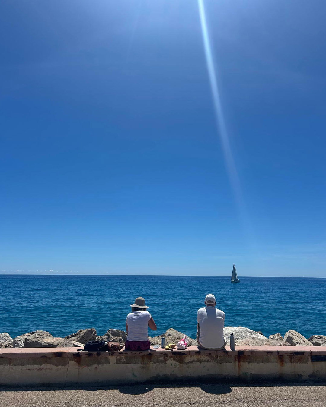 Two people sitting on a sea wall facing a deep blue sea and distant sailboat under a clear sky and sunbeam