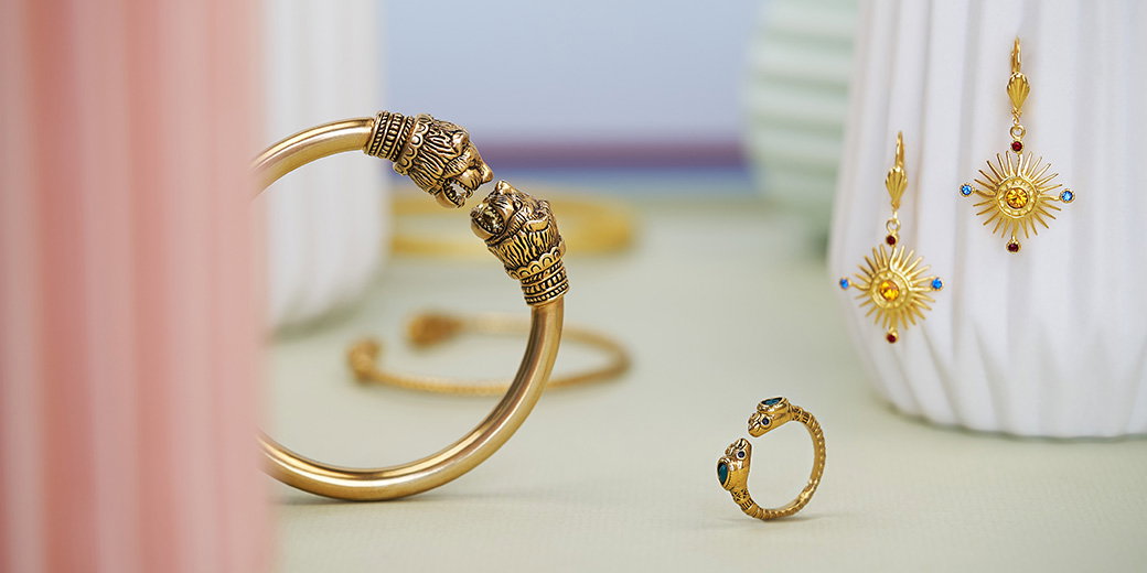 Gold jewellery including Holy Thorn Reliquary earrings, a lion head bracelet and a green agate snake ring displayed against a soft pastel background with ceramic origami forms. - Jewellery Collection.