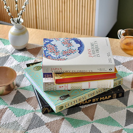 Stack of illustrated hardback books including Cultural Treasures, History of the World: Map by Map and Japan: A History in Objects on a coffee table with a vase and tea. - Books Collection.