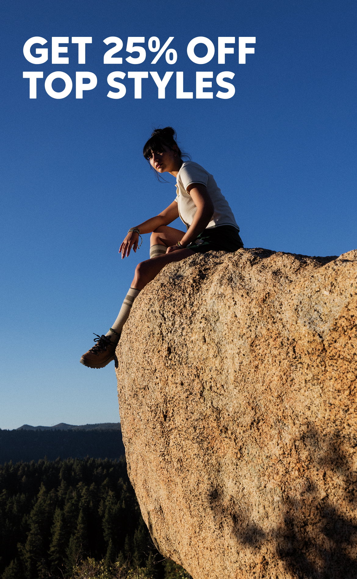 A woman sitting on a rock, wearing a white shirt, green shorts, and hiking boots with text: GET 25% OFF TOP STYLES 
