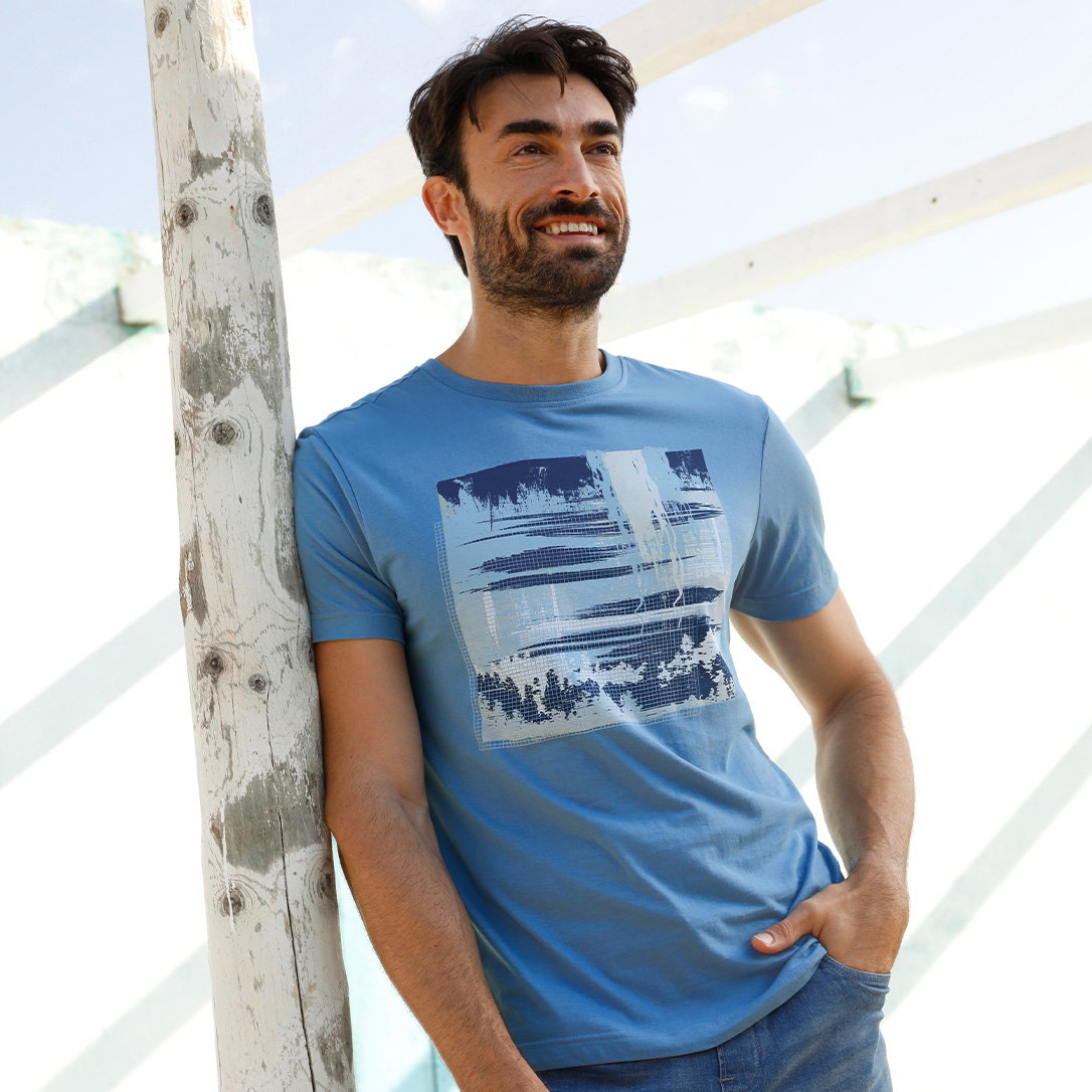 Ein männliches Model trägt ein blaues T-Shirt mit abstraktem Print und lehnt an einem weißen Baumstamm im Freien.