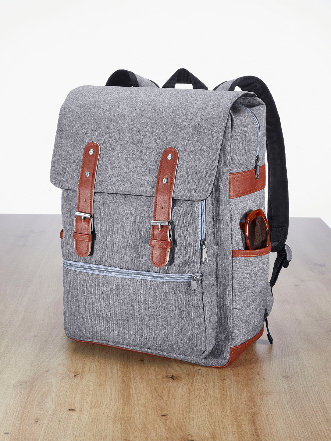 Ein grauer Rucksack mit braunen Details, mehreren Reißverschlusstaschen und einem seitlichen Fach mit Sonnenbrille, steht auf einem Holzboden.