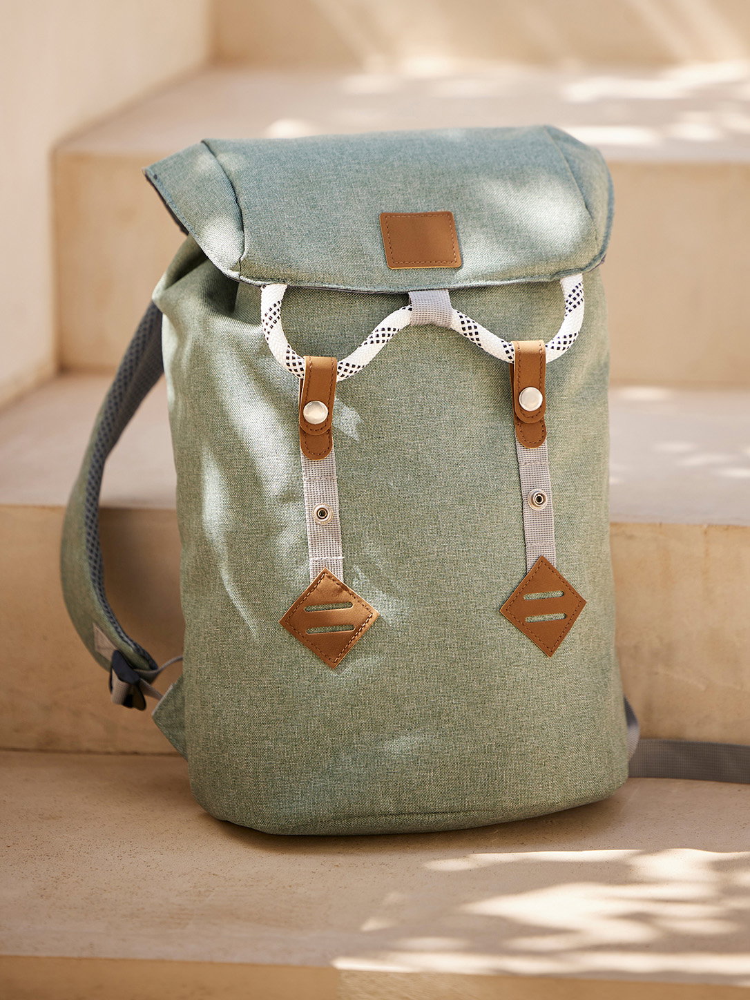 Ein petrolfarbener Rucksack mit braunen Details und verstellbaren Schultergurten, steht auf hellen Stufen im Sonnenlicht.
