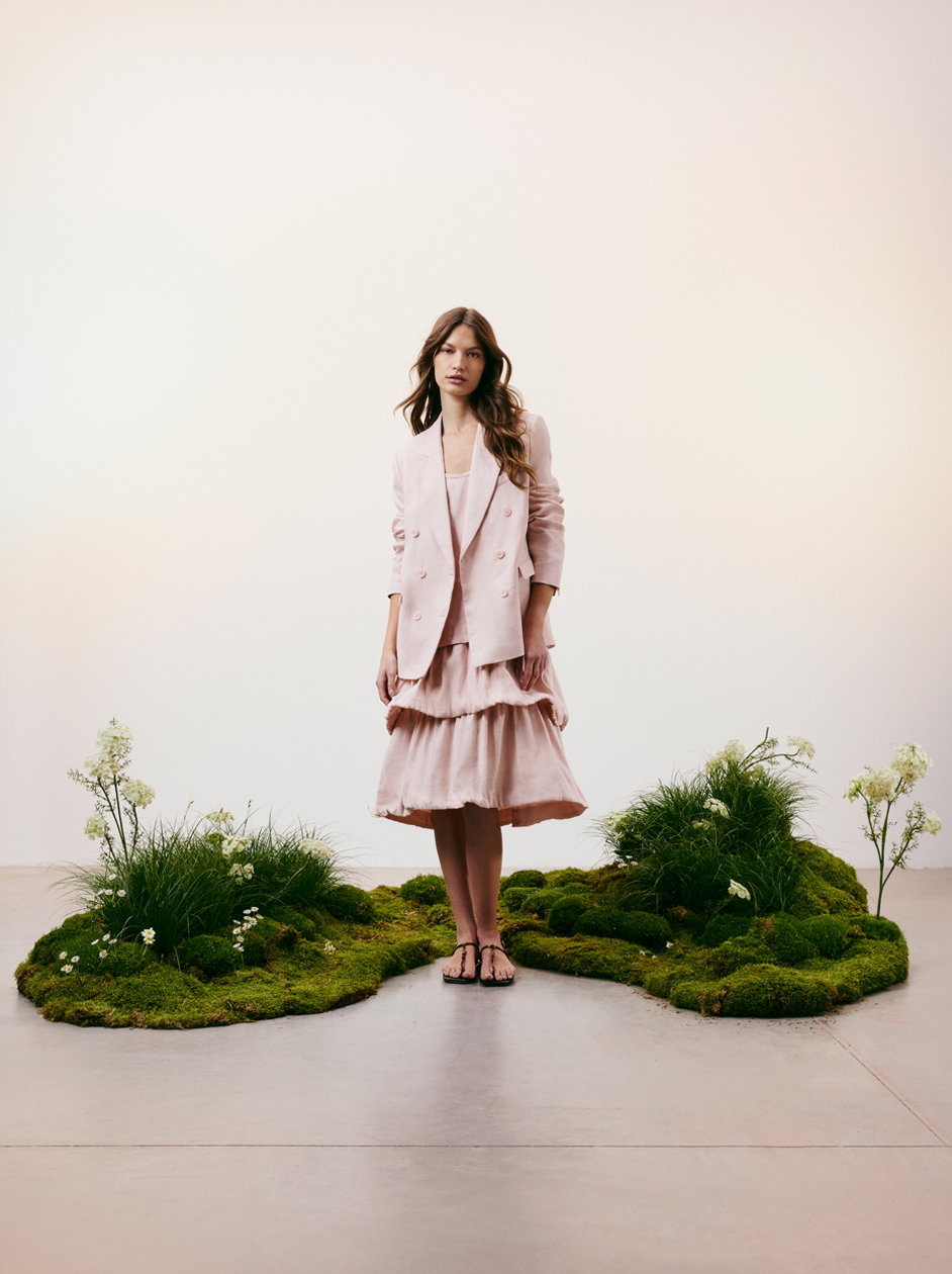 Ein Model von DOROTHEE SCHUMACHER präsentiert ein elegantes Ensemble in zartem Rosé, bestehend aus einem doppelreihigen Blazer und einem luftigen Stufenrock. Sie steht auf einer kunstvoll arrangierten Moosinsel mit Gräsern und kleinen weißen Blüten vor einem hellen, minimalistischen Hintergrund.