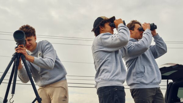 Three teenagers wearing gray sweatshirts birdwatch. On the left, one boy is looking through a scope, while the two others on the right are looking through binoculars. 