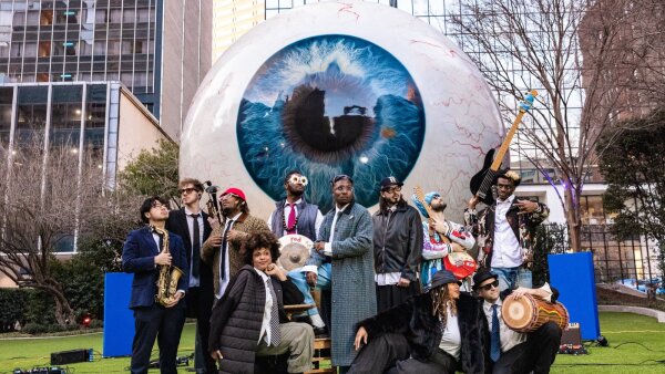Cure For Paranoia, a musical collective of at least 11 people, stands in front of a large eyeball that has a blue eye color. 