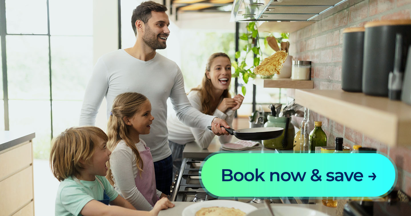 A family smiles as a parent flips a pancake over the stove in a bright kitchen, with a 'Book now & save' button displayed in front