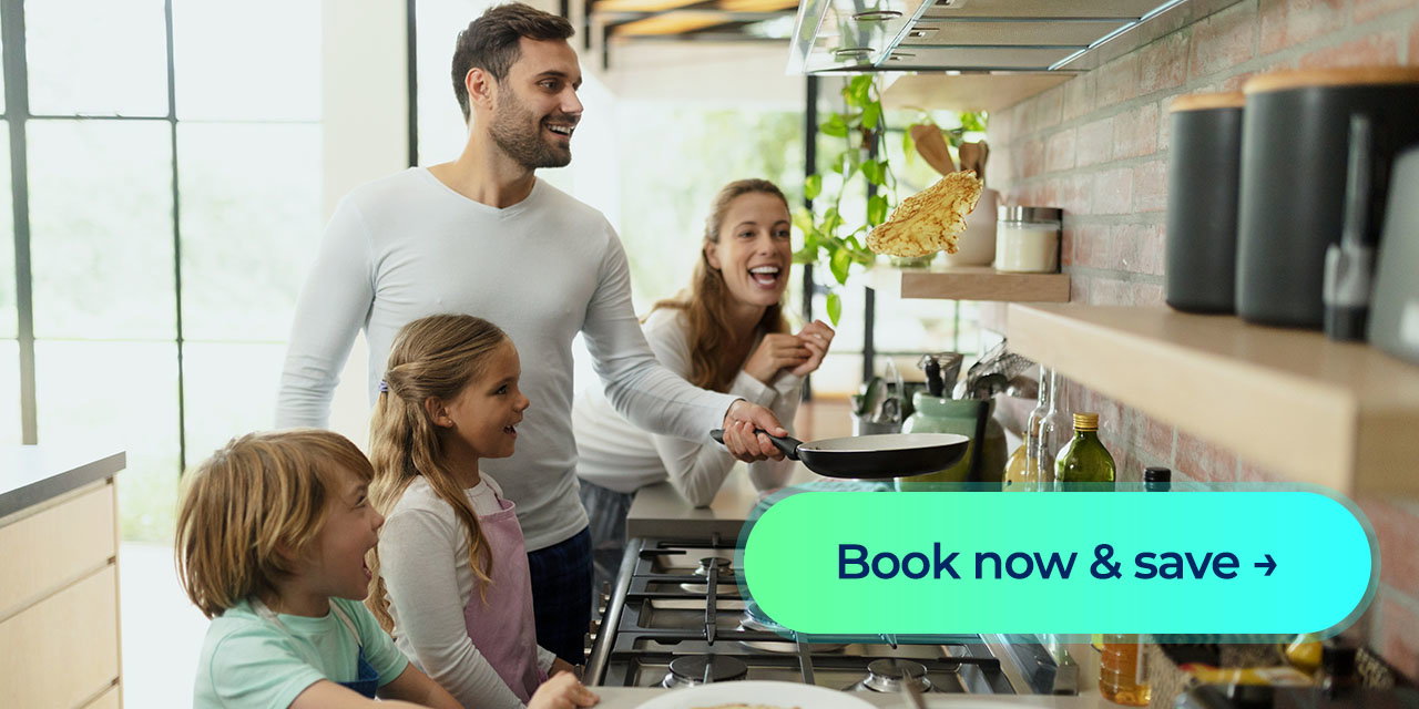 A family smiles as a parent flips a pancake over the stove in a bright kitchen, with a 'Book now & save' button displayed in front