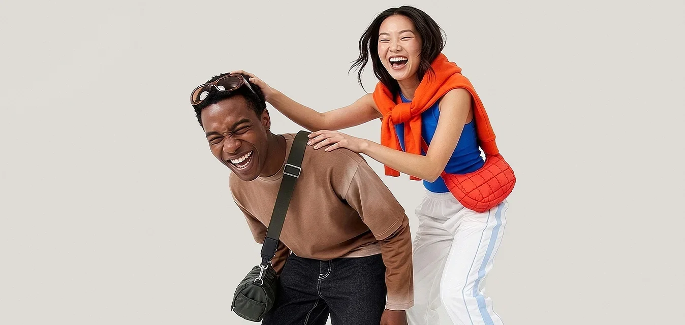 Jonge man lacht, voorover gebogen met crossbodytas, vrouw achter hem glimlacht, handen op zijn schouders, draagt een oranje trui en sandalen.