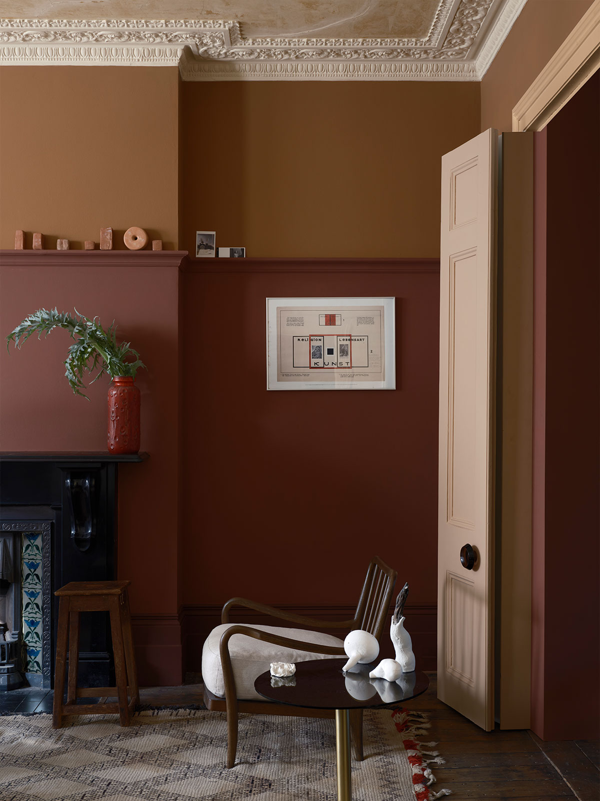 A cocooning living room decorated in a mix of earthy colours. Dark brown skirting and walls in deep red and burnt orange are lifted by a soft salmon pink ceiling and matching door. Simple furniture, a fireplace and soft textures balance the scheme.