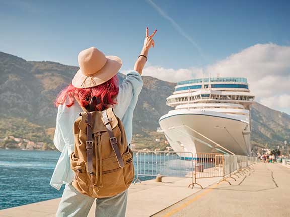 Frau mit roten Haaren, Hut und Rucksack läuft an einem sonnigen Tag auf einem Steg zu einem großen Kreuzfahrtschiff zu.