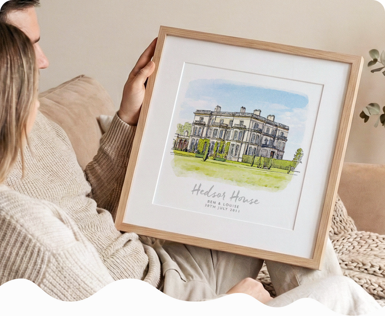 A woman holding a framed watercolour sketch of a grand country house, personalised with the names "Mr & Mrs Harrison" and their wedding date