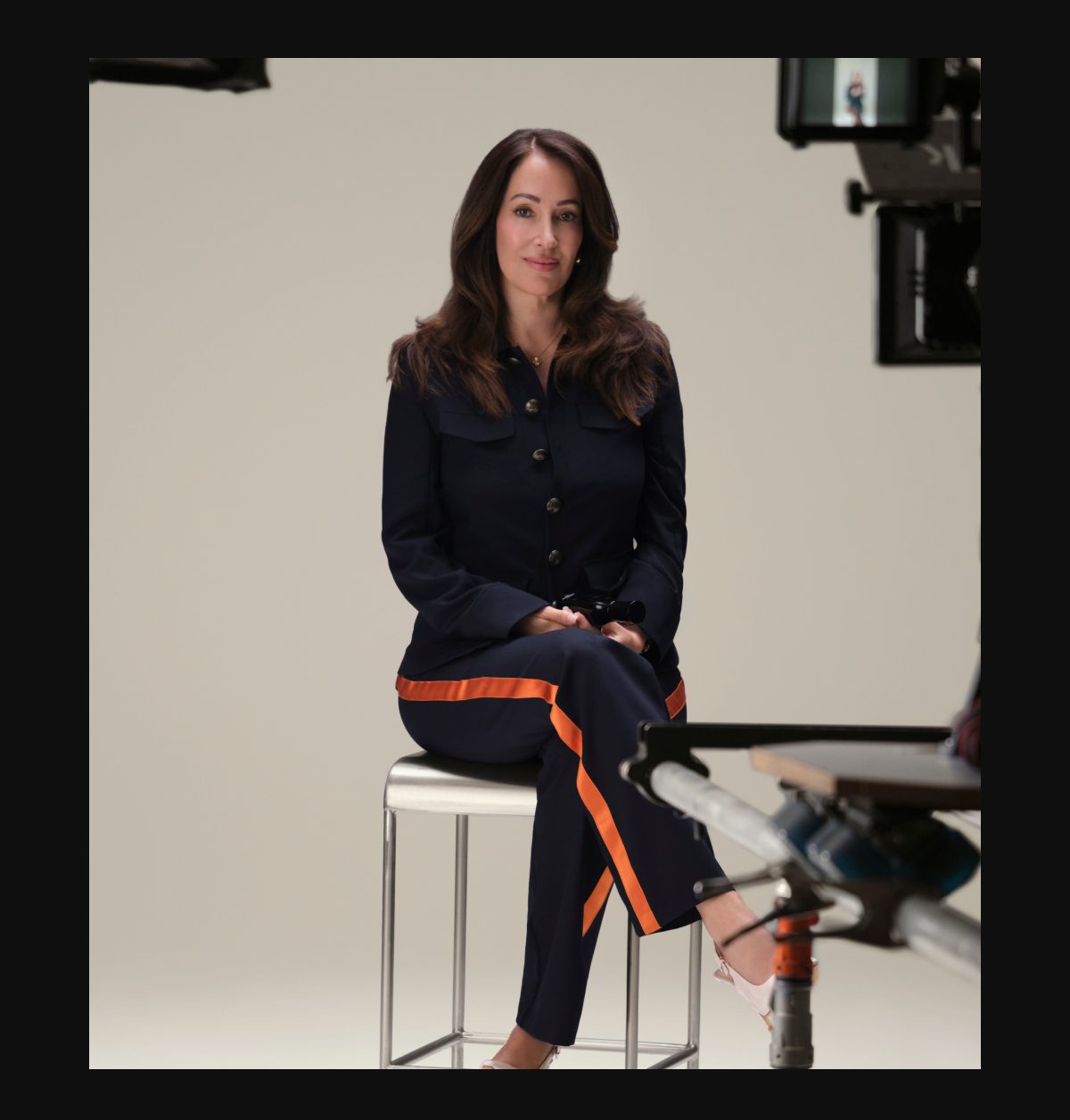 Angela Caglia sitting on a stool, wearing a navy outfit with orange stripes, in a studio setting.