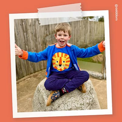 A boy wearing a blue tiger top and Start-Rite Tide camo canvas, sitting on a rock with his arms outstretched.