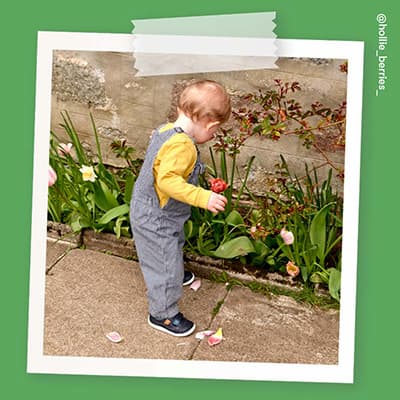 A toddler wearing a yellow top and blue dungarees with Start-Rite Playhouse navy shoes, picking flowers in the garden.
