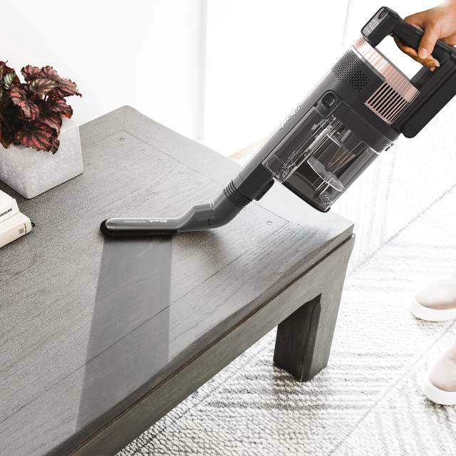 A person uses a Shark vacuum cleaner to clean a dark wooden table. A plant is on the table.