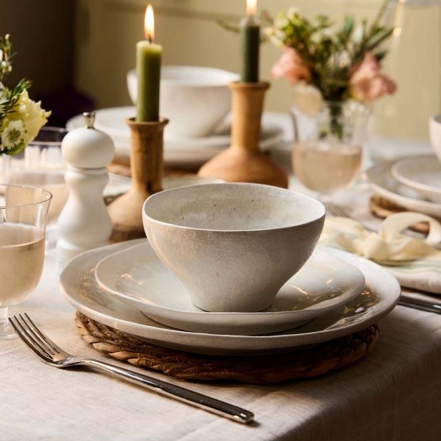 Elegant table setting with a bowl, plates, wine glass, candles, and flowers on a white tablecloth.