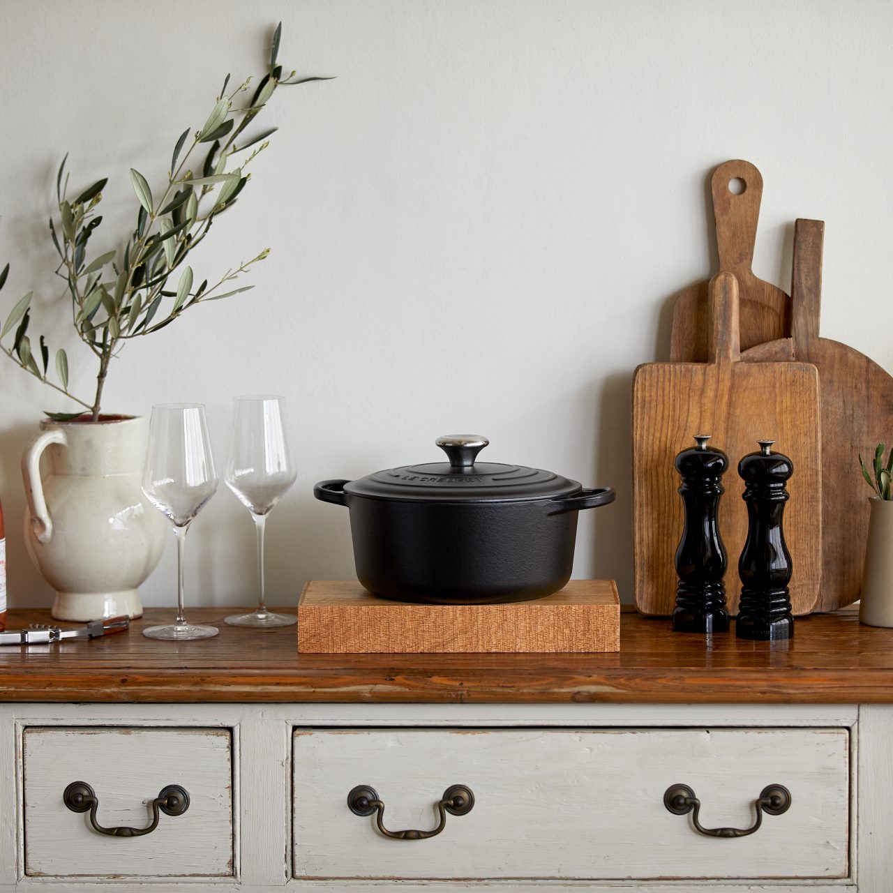 A rustic kitchen setup with a black pot on a wooden pedestal, cutting boards, wine glasses, and a vase with leaves.