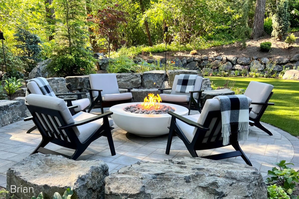 Breeze chairs with light grey cushions arranged around a round fire pit on a stone patio with landscaped greenery.