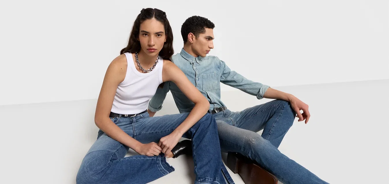 Twee personen zitten op een witte ondergrond. De een draagt een witte tanktop en blauwe jeans, de ander een licht denim shirt en donkere jeans.
