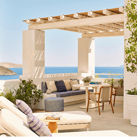 Shaded white stucco terrace with cushioned lounge seating, a round dining table and chairs, and two sun loungers overlooking the coastline and blue sea.