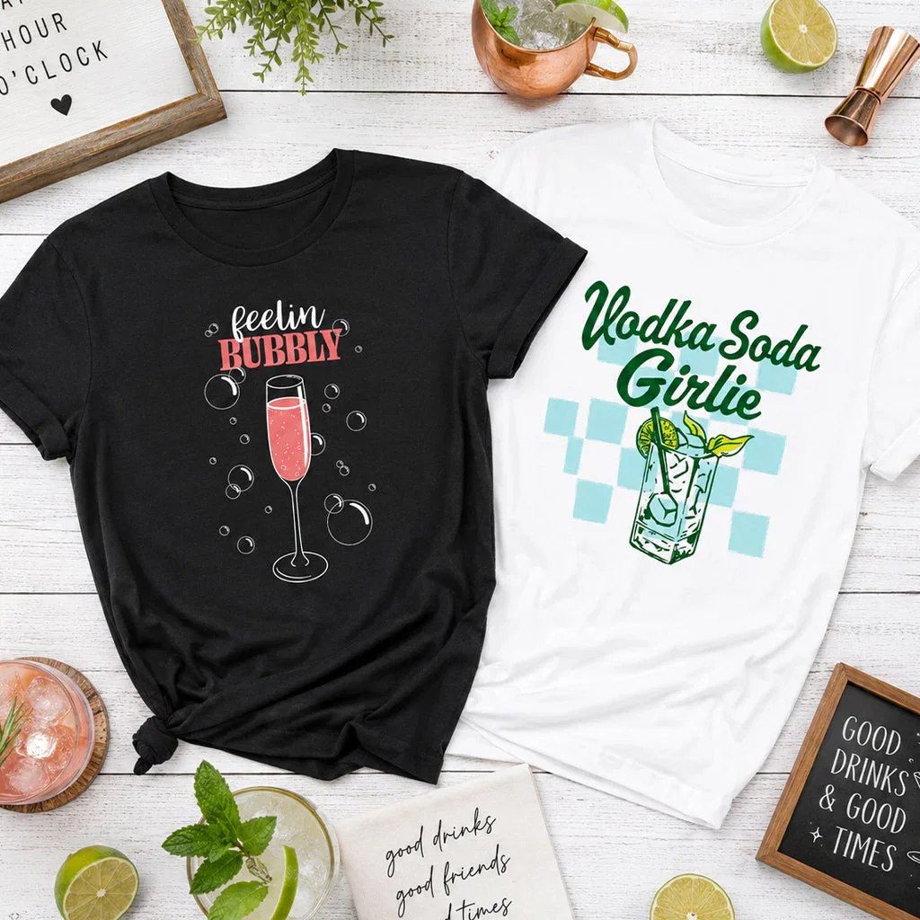 Two graphic t-shirts displayed on white wood with cocktail props and decorative signs promoting the Happy Hour O'Clock collection.
