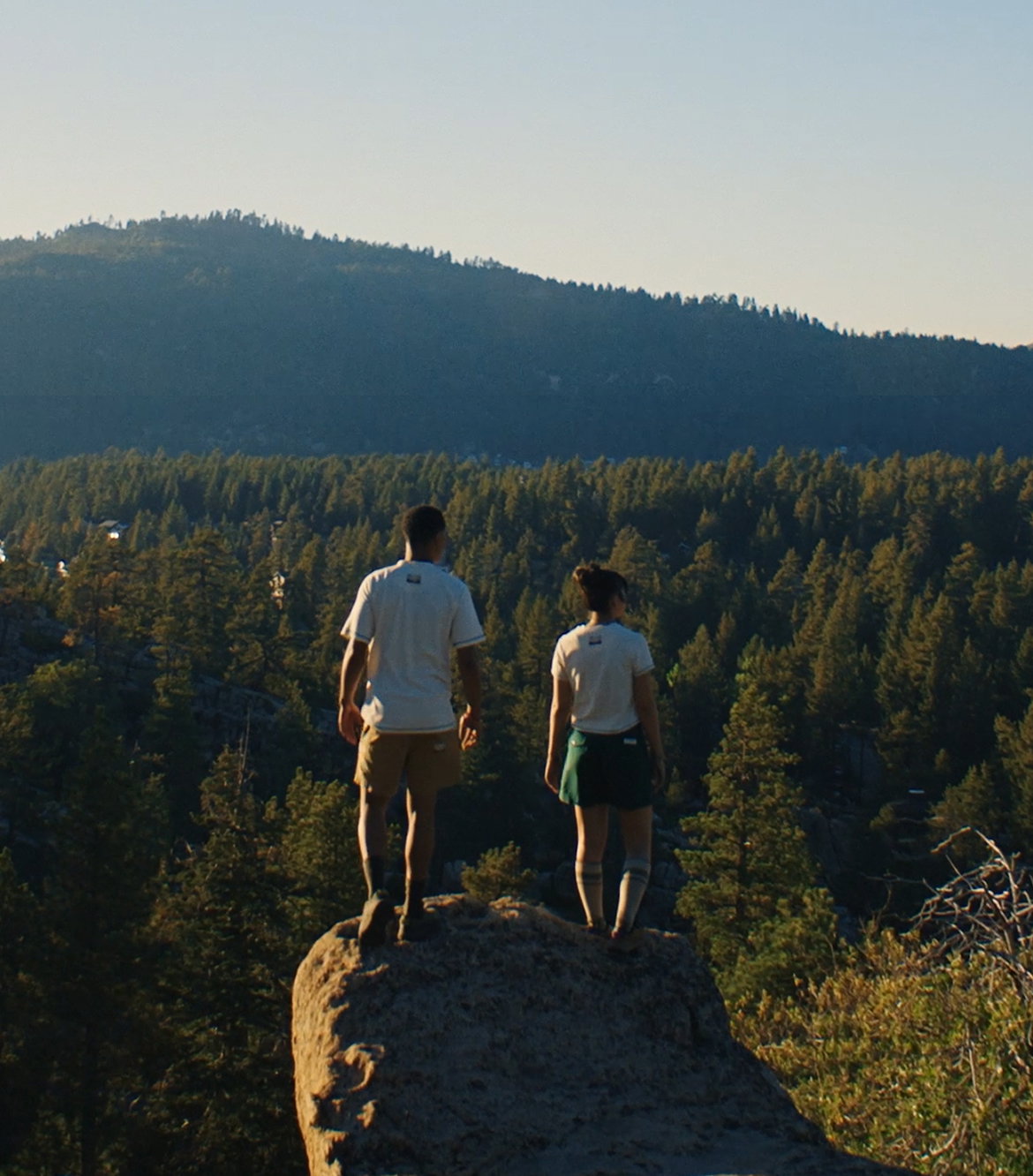 A man and a woman standing on a rock overlooking a forest, wearing white shirts, khaki bottoms, and hiking boots. 