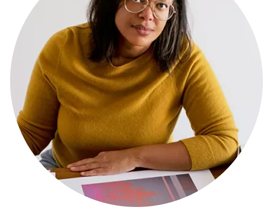 Person seated at a table with artwork, wearing glasses and a mustard colored top.
