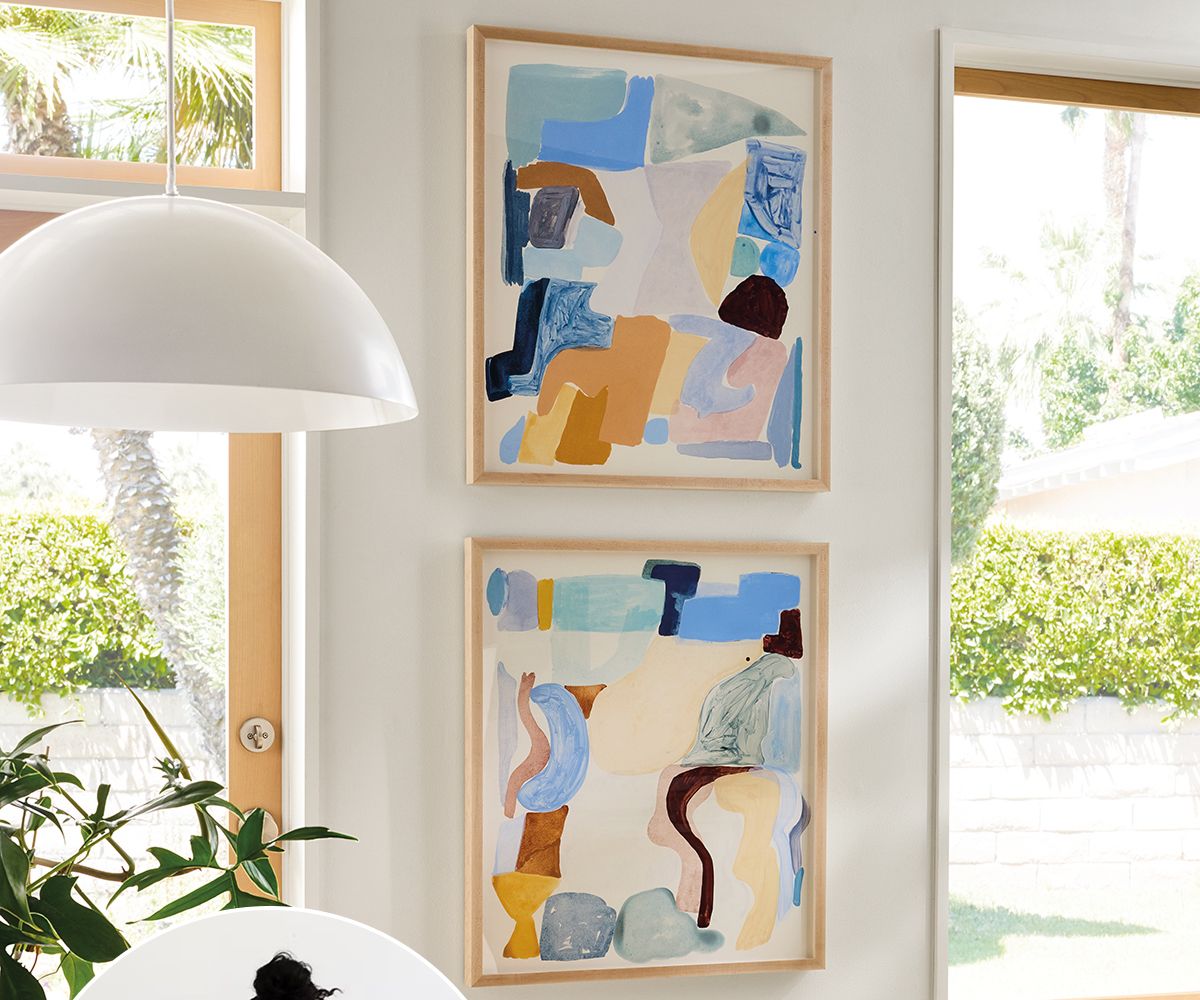 Two framed abstract artworks with blue, tan, and cream shapes hang between windows beside a white pendant light.