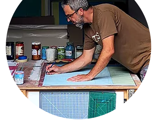 Artist sketching at a table with rulers and materials spread across a work surface.
